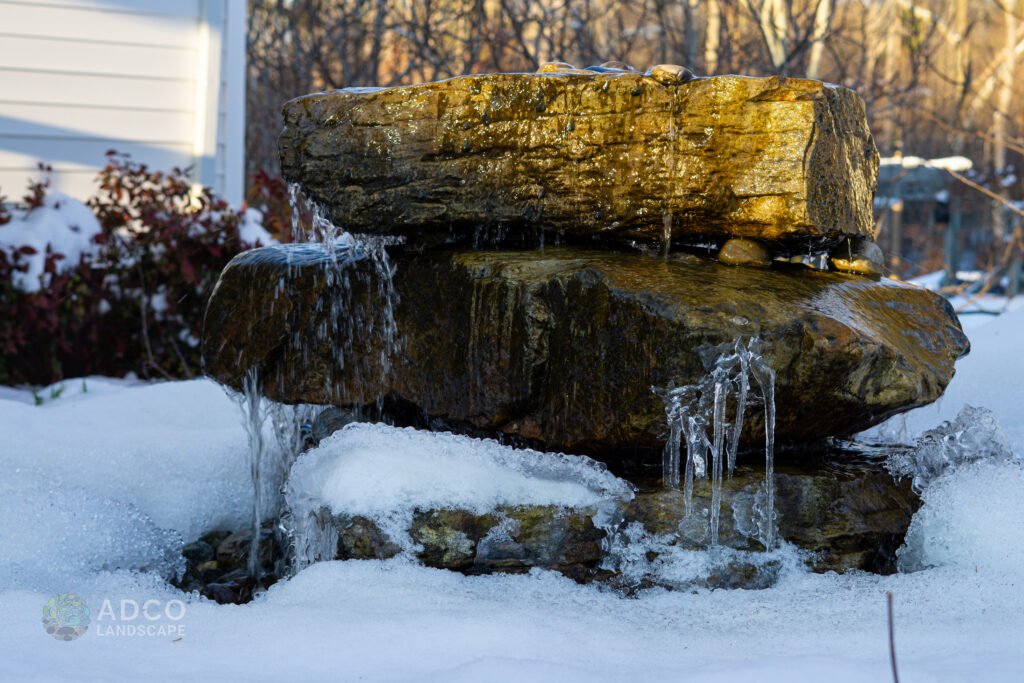 A Custom Natural Boulder Water Feature with red shrubs in the background in the snow.