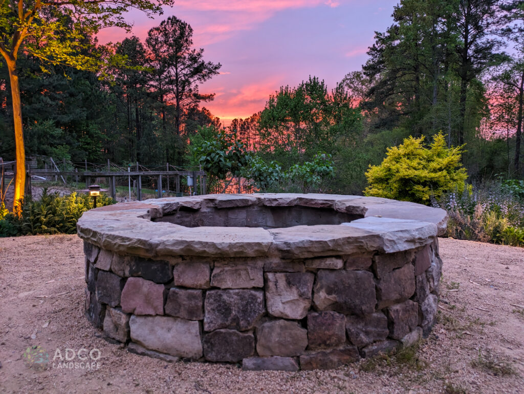 A stone fire pit with flowers, trees, and a sunset in the background.